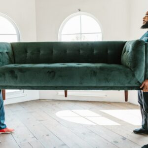 Two men lifting a green sofa in a well-lit room with arched windows.